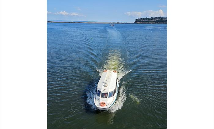 A white Aquabus passenger ferry boat cruising through the blue water of Cardiff Bay, leaving a wake behind it, with the coastline visible in the dista